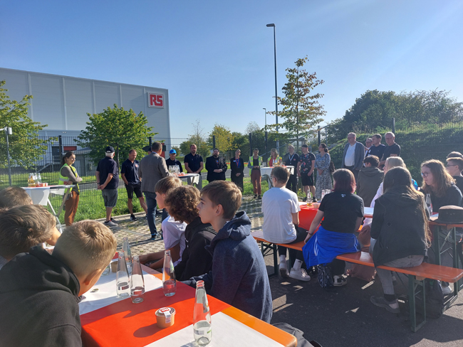 Schüler auf Bänken im Vordergrund, hinten stehen Leute an Tischen, Sonnenschein