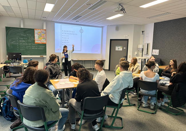 Schüler im Klassenraum, Lehrerin gestikulierend vor Beamer und Tafelbild