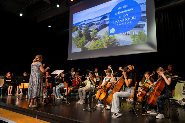 Bühne mit Orchester aus Schülern, davor dirigiert eine Lehrerin, im Hintergrund Beamerbild mit Willkommensgruß