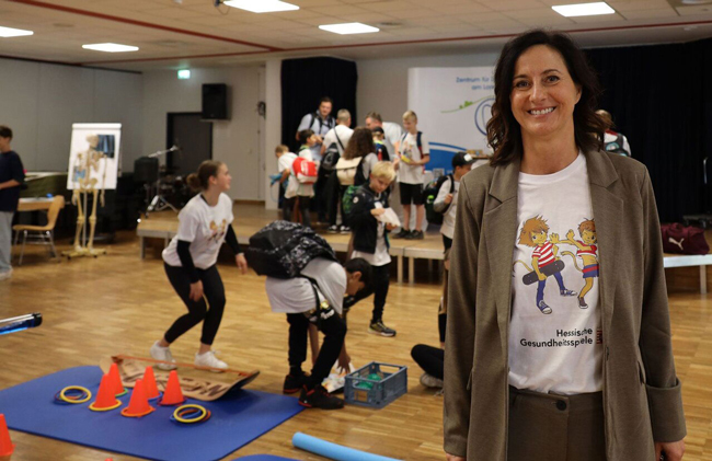 Schulleiterin im Vordergrund, im Hintergrund Aula mit Stationen zum Gesundheitslernen