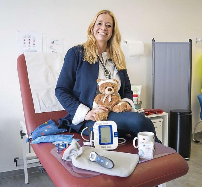 Corinna Knobel sitzt auf Behandlungsliege lächelnd mit Teddy in den Händen, um sich herum liegen Blutdruckmessgerät, Fieberthermometer, Tasse, Wärmflasche und andere Hilfsmittel