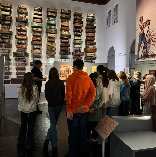 Schüler stehen vor einer großen Wand mit verschiedenen alten Koffern, Museumsführer spricht zu ihnen