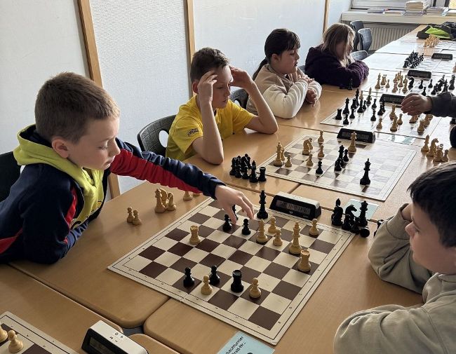 Jungs sitzen sich gegenüber an Tischen mit Schachbrettern zwischen sich, einige Schachfiguren stehen neben den Brettern, Schüler wirken konzentriert