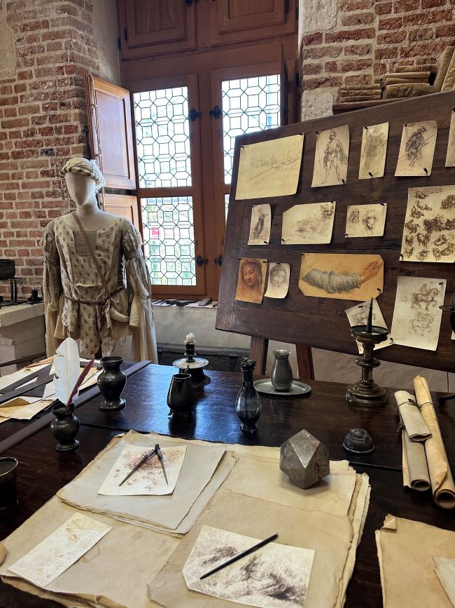 Blick in ein Museum mit Schriftrollen, Skizzen und alten Gefäßen auf einem Tisch und einer Statue vor einer Backsteinwand