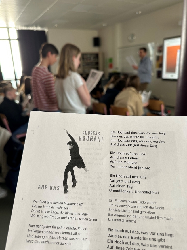 Im Vordergrund ein Liedtext, im Hintergrund stehen Schüler mit Texten in der Hand zusammen