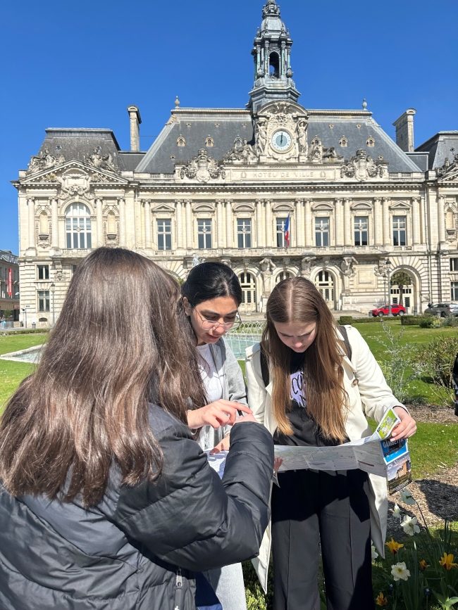 drei Schülerinnen blicken auf einen Plan, im Hintergrund ein Schloss bei blauem Himmel