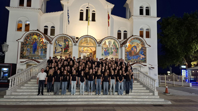 Chorgruppe mit schwarzen Poloshirts vor einer Kirche mit bunten Glasfenstern am späten Abend