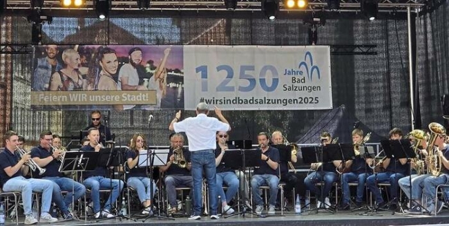 Bläsergruppe musiziert auf einer Bühne, dirigiert von einem Mann mit weißem Hemd im Vordergrund