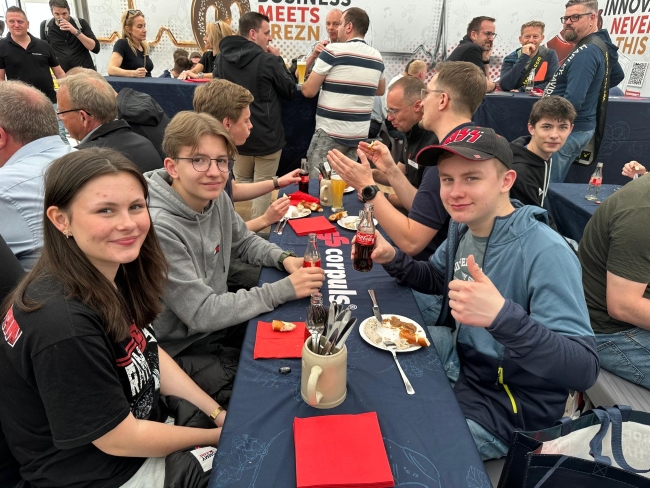 Schüler sitzen an Bierzeltgarnitur und essen, lächeln in Kamera
