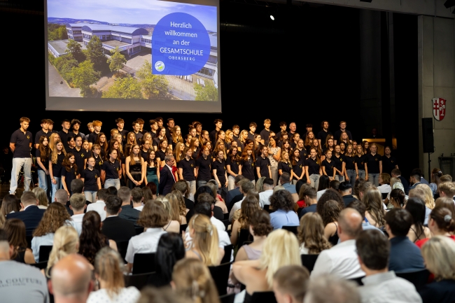 Chor vor Publikum auf Bühne, im Hintergrund großes Beamerbild der Schule