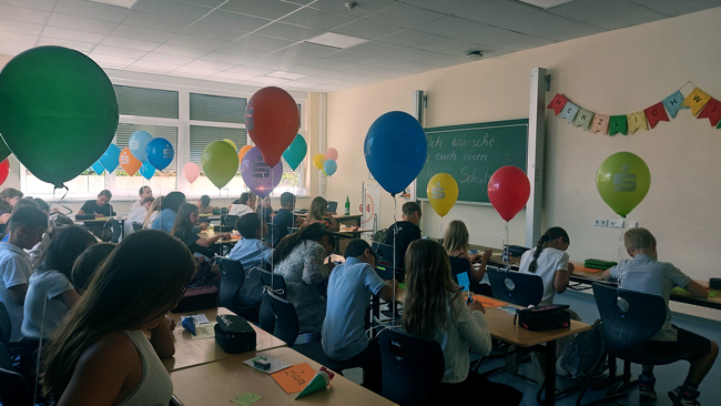 Blick in Klassenraum voller bunter Luftballons, Kinder sitzen an Tischen und schreiben etwas auf
