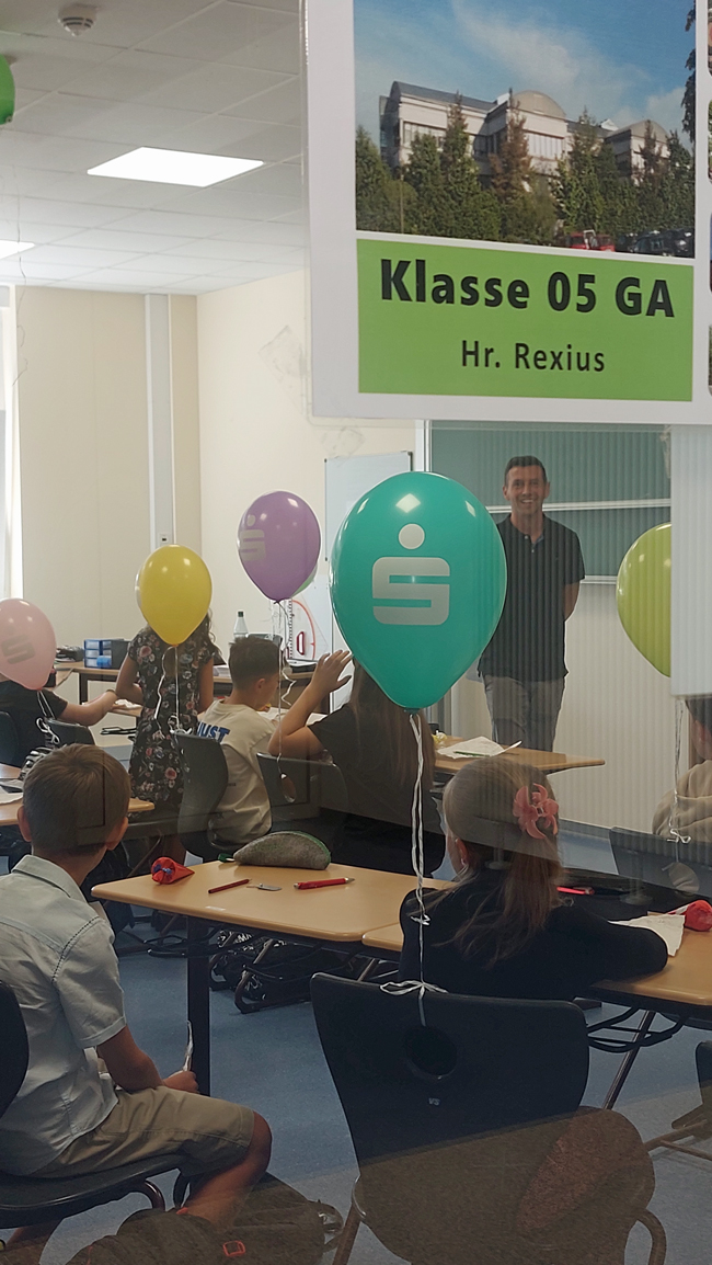 Blick in andere Klasse durch Tür, an der ein Schild hängt mit der Aufschrift "Klasse 05 GA Herr Rexius", Lehrer im Hintergrund lächelnd zu sehen