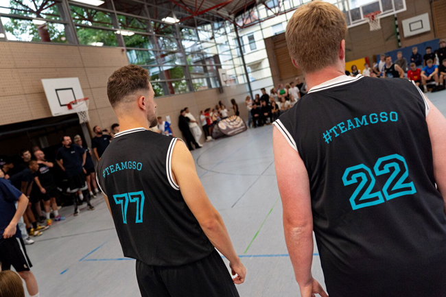 Zwei Lehrer von hinten mit der Trikotaufschrift "teamGSO" auf das Spielfeld blickend