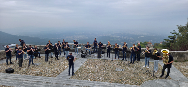 Musiker mit Blechblasinstrumenten auf einem hoch gelegenen gepflasterten Platz, umgeben von einer Mauer, hinter der man weit ins Land blicken kann