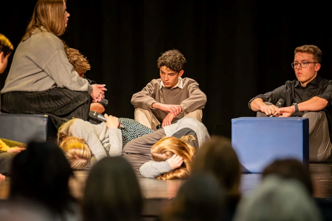 Schüler auf der Bühne bei einem Schauspiel, einige liegen schlafend, andere sitzen oder hocken und starren ernst ins Leere