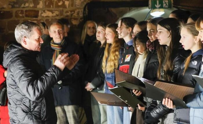 Chorleiter dirigiert vor Schülern mit Notenblättern in der Hand und warm gekleidet singend im Freien