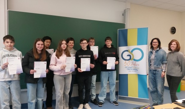 Schüler mit Urkunden vor einer Tafel in einem gr0ßen Klassenraum blicken lächelnd in die Kamera